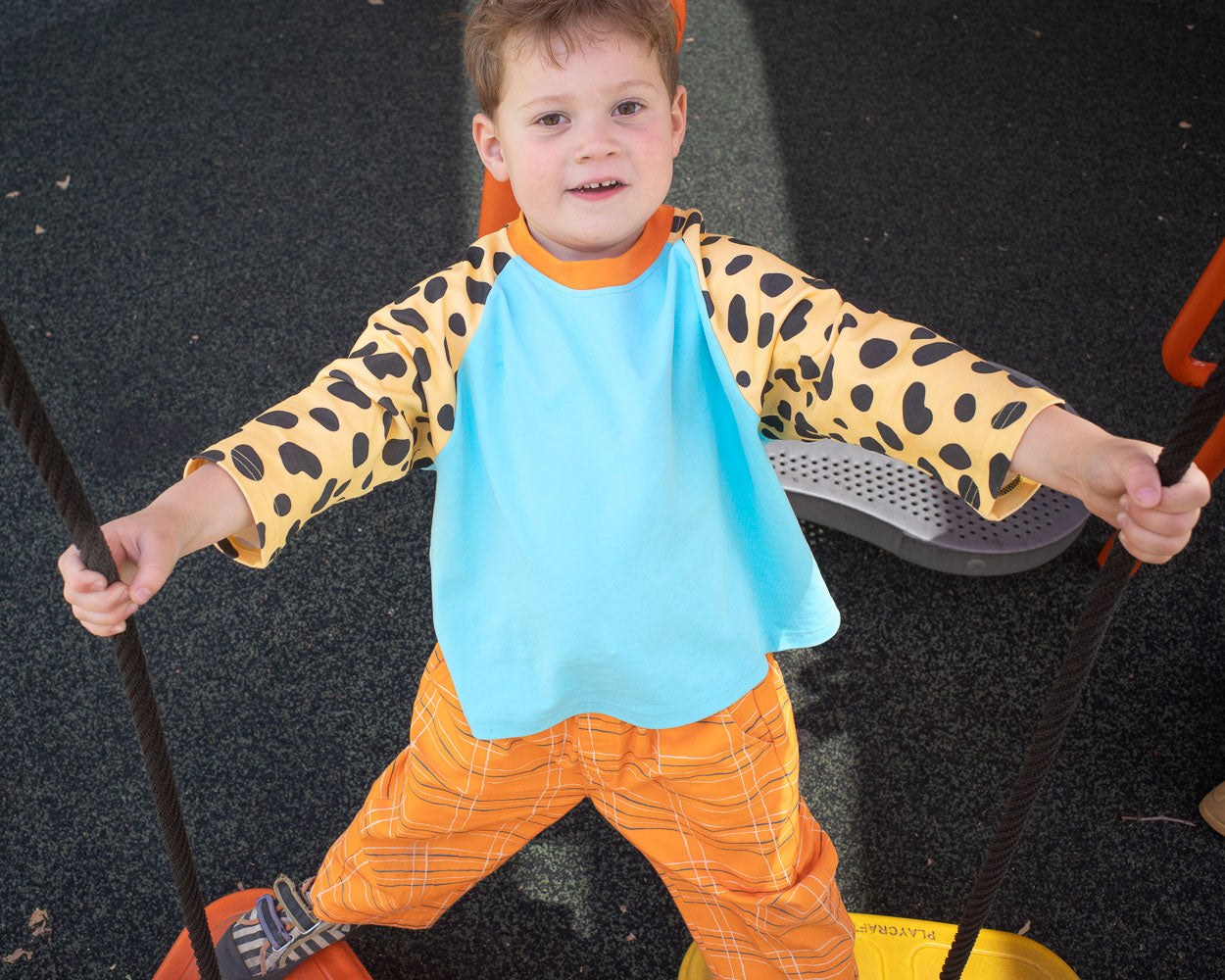 Child wearing a blue raglan t-shirt with yellow and black spotted sleeves and orange pants on a dark background.