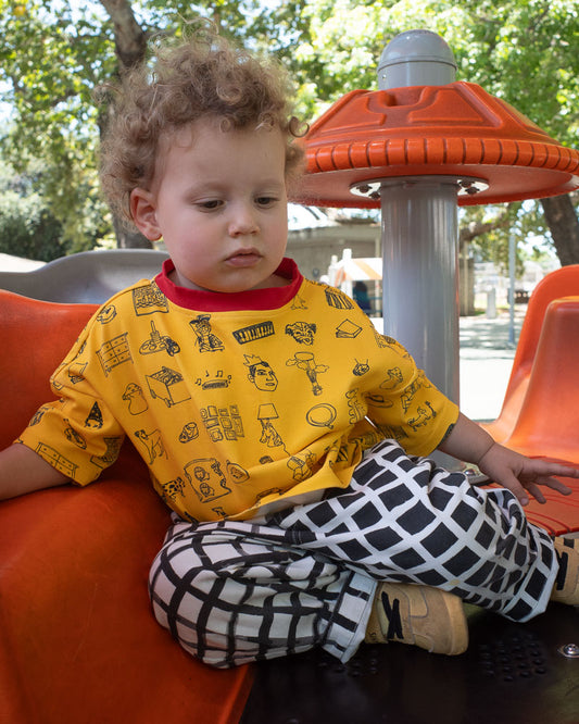 Child sitting on an orange playground structure wearing a yellow shirt with black doodle patterns and black and white checkered pants.