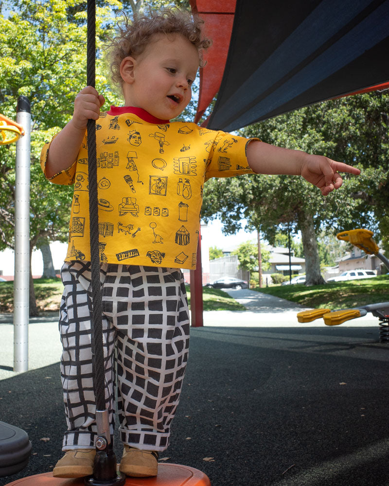 Child in a yellow shirt with black doodle print and check pants standing on a playground structure, holding onto a rope.