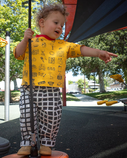 Child in a yellow shirt with black doodle print and check pants standing on a playground structure, holding onto a rope.
