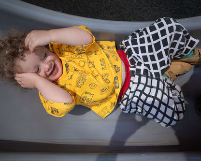 Child wearing a yellow shirt with doodle patterns and a black and white checkered outfit, lying on a slide.