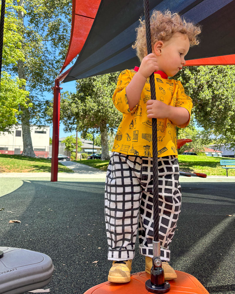Child in a yellow shirt and checkered pants standing on a playground equipment.