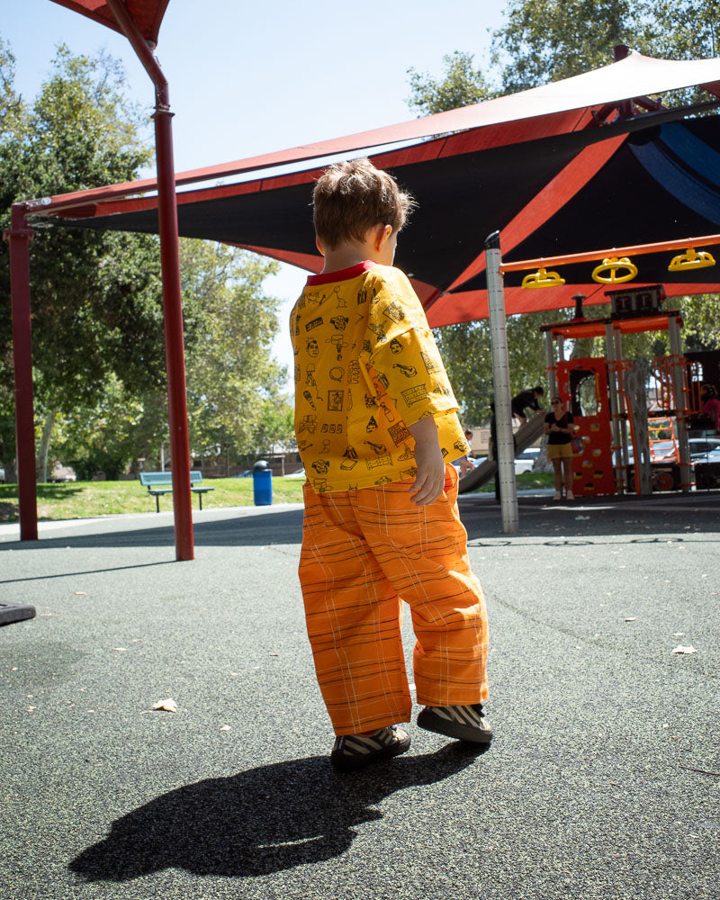 Child in a yellow shirt and orange plaid pants standing on a playground